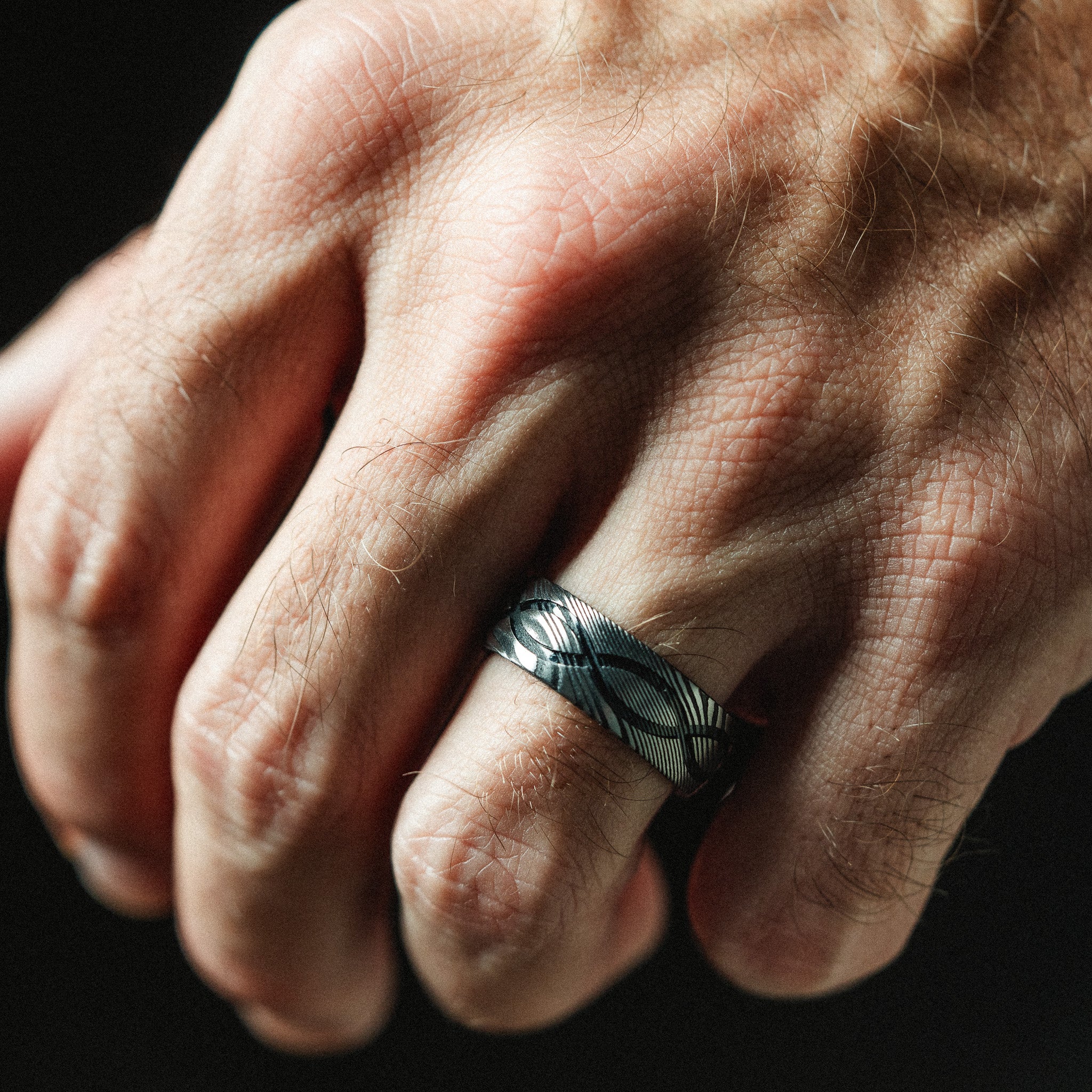 Close-up of a hand wearing a silver ring with a black band against a dark background