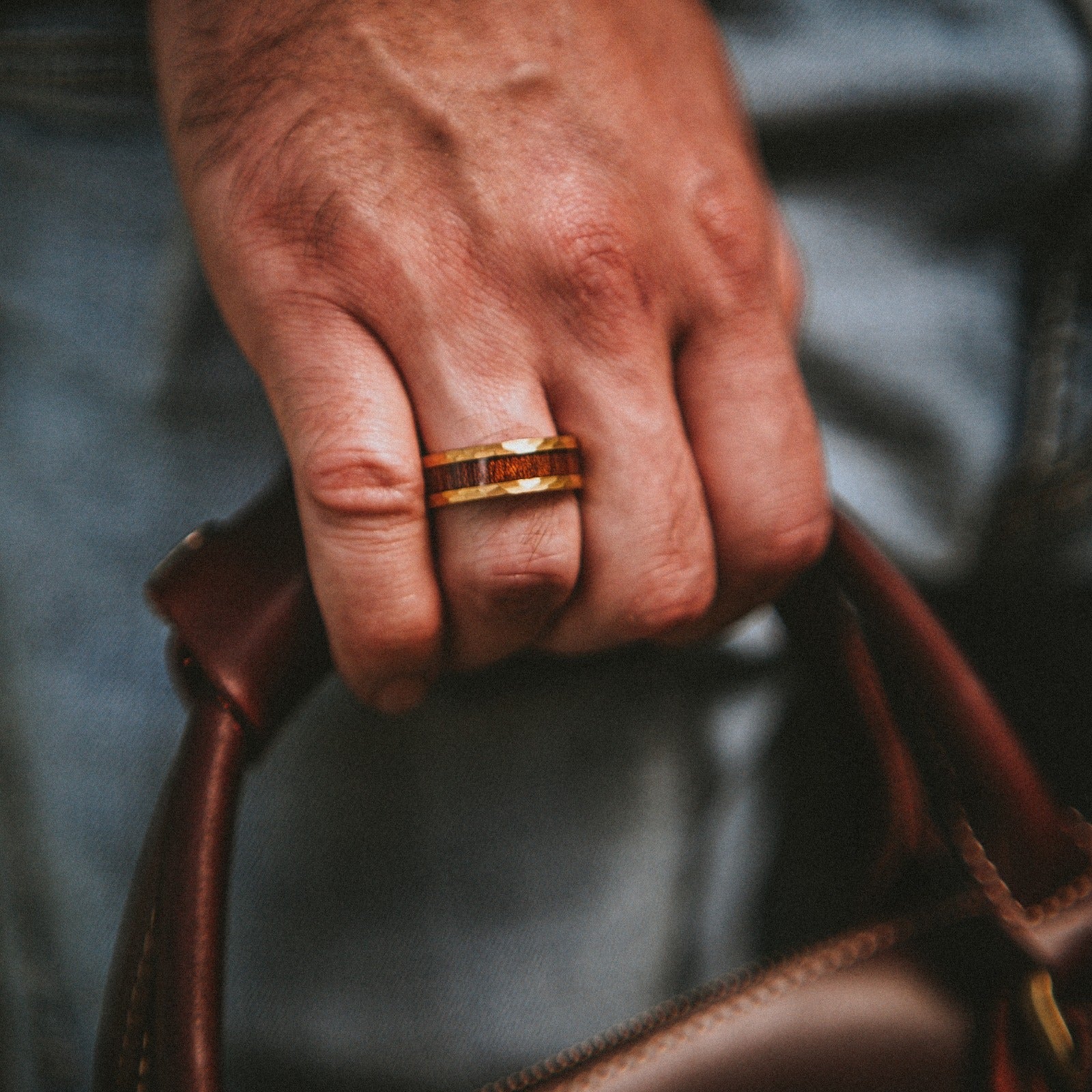 Hand wearing a gold ring holding a brown leather bag against a blurred background