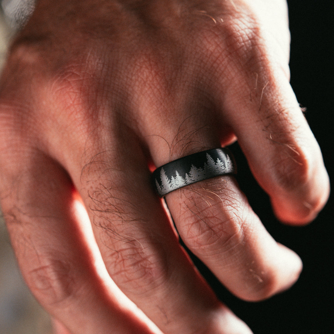 Close-up of a hand wearing a black ring with a forest design on a dark background