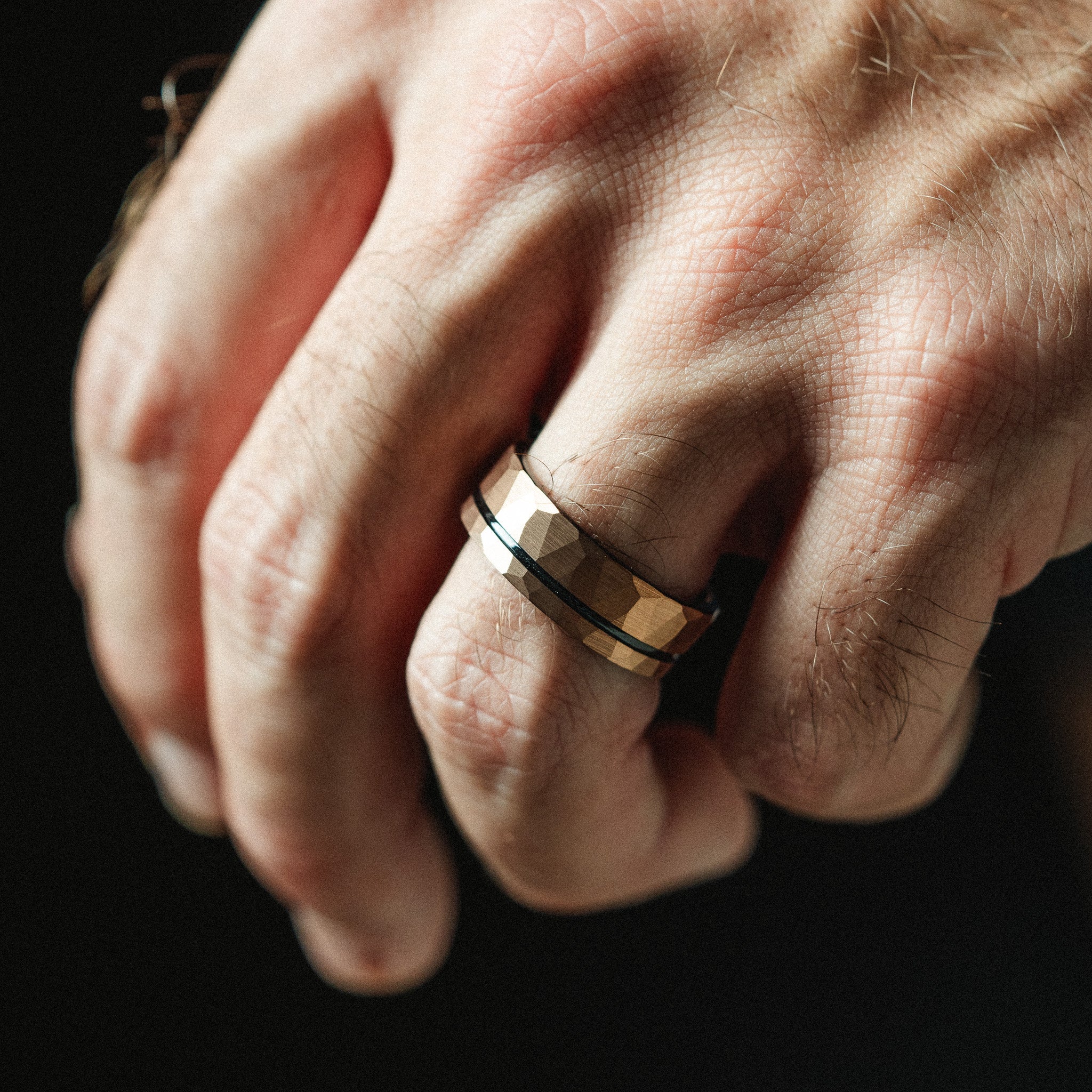 Close-up of a hand wearing a gold ring with a black band on a dark background